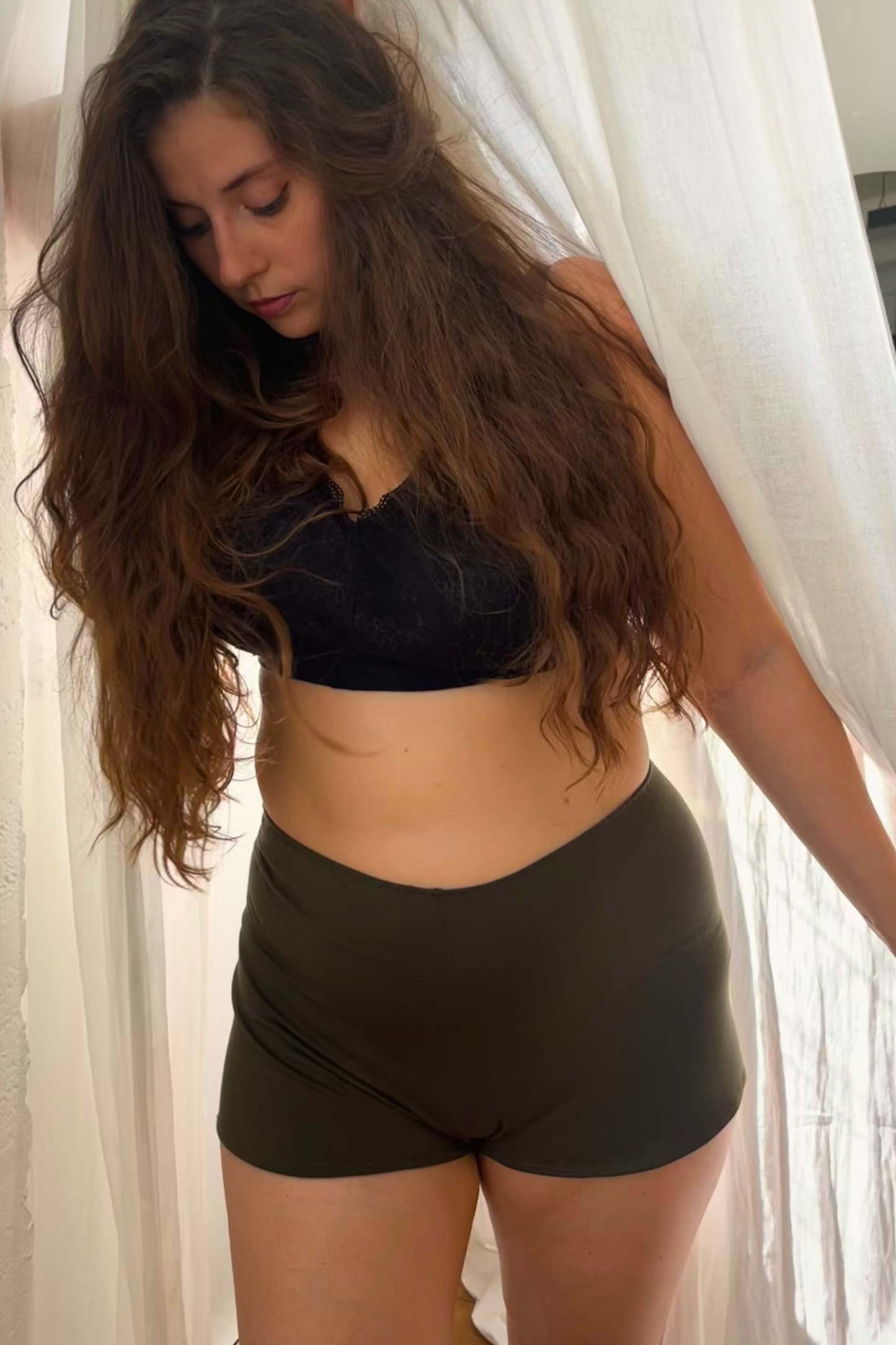 Woman wearing a black bra and high-waisted shorts against a white curtain background