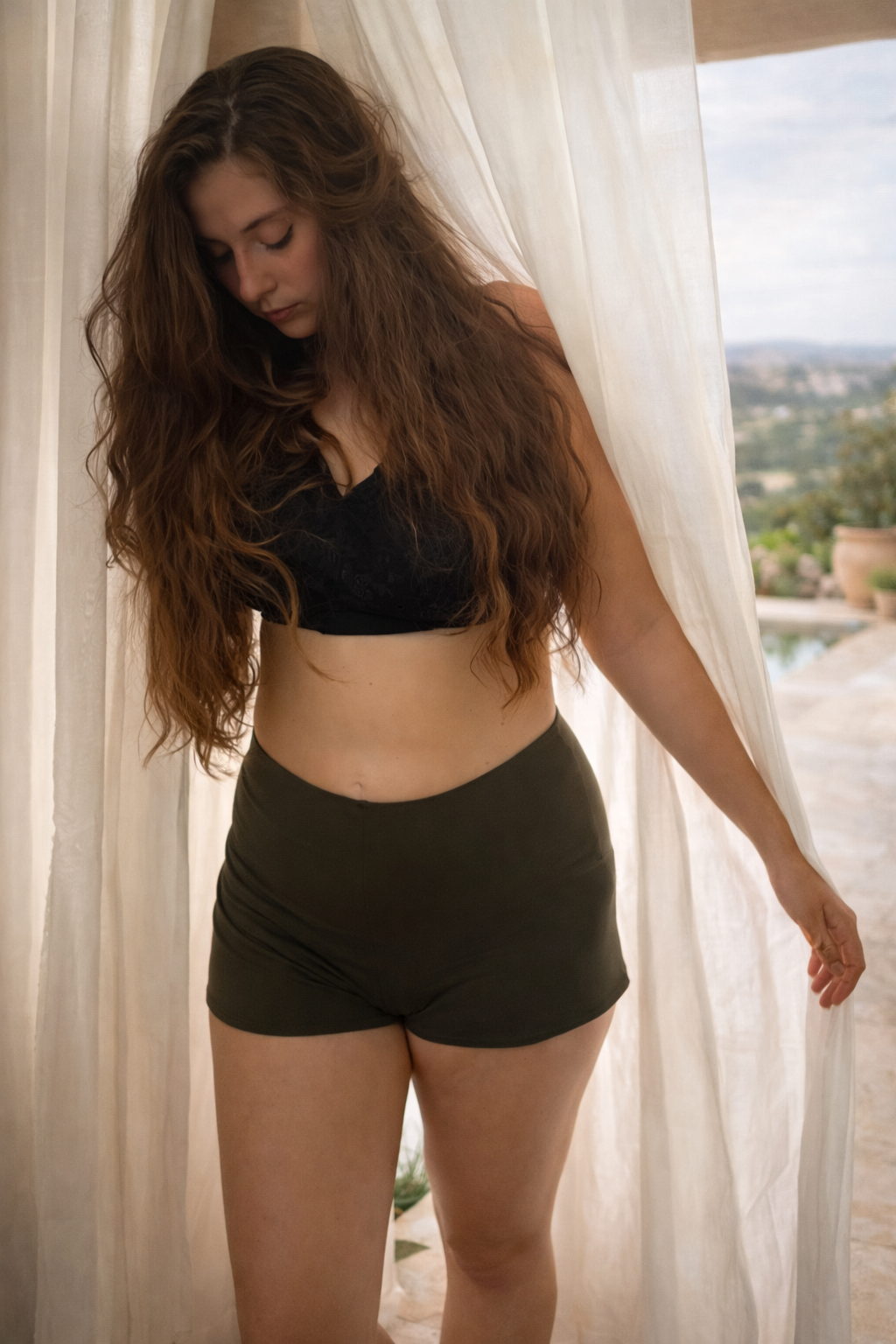Woman wearing a black bikini top and green shorts standing in front of sheer curtains with a scenic background.