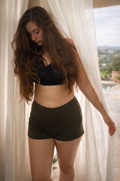 Woman wearing a black bikini top and green shorts standing in front of sheer curtains with a scenic background.