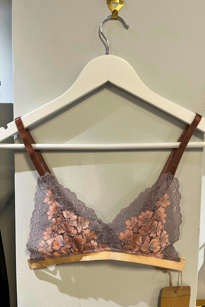 hand-sewn metallic rose lace bralette hanging on a white hanger, showing the dual-tone lace, wide straps and triangle nipple-cover lining, photographed against a soft studio background.