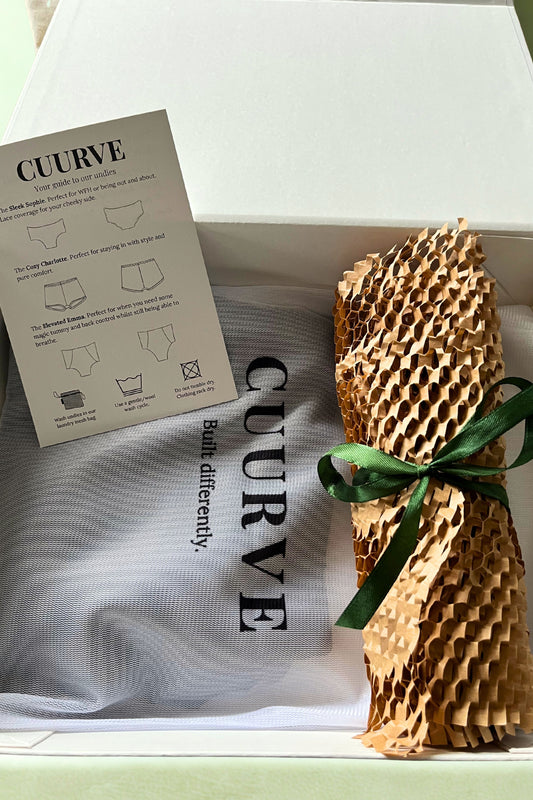 Open Cuurve gift box showing Cosy Charlotte pajama shorts in Dark Olive with packaging, care card, and green ribbon — sustainable slow-fashion gift set.