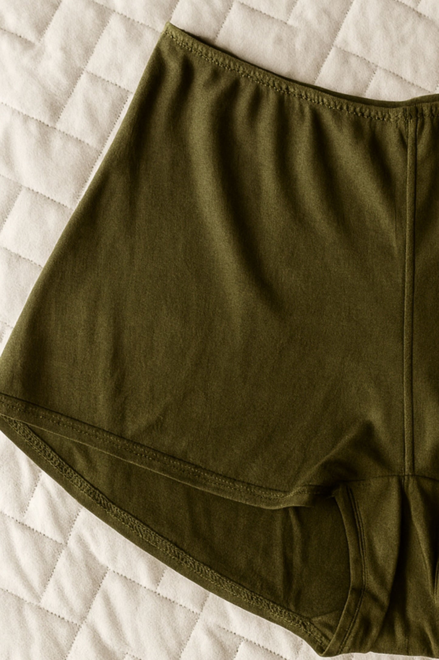 Flat lay of Cuurve Cosy Charlotte pajama shorts in Dark Olive on quilted bedding — minimalist slow-fashion design made in England.